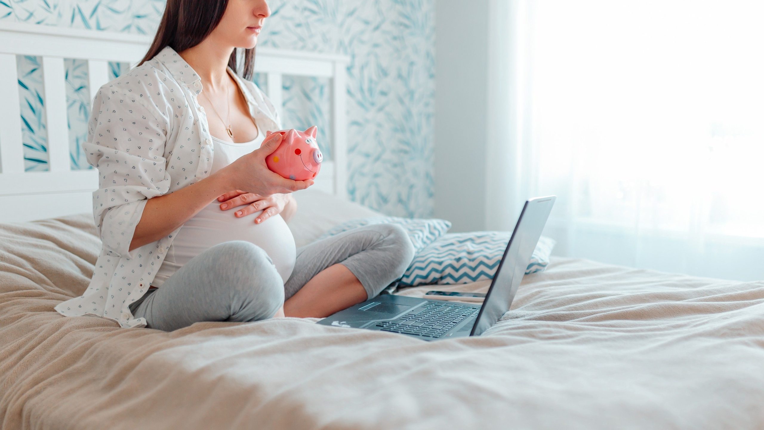Pregnant woman sitting on bed with laptop, while holding a piggy bank and financially preparing for a baby