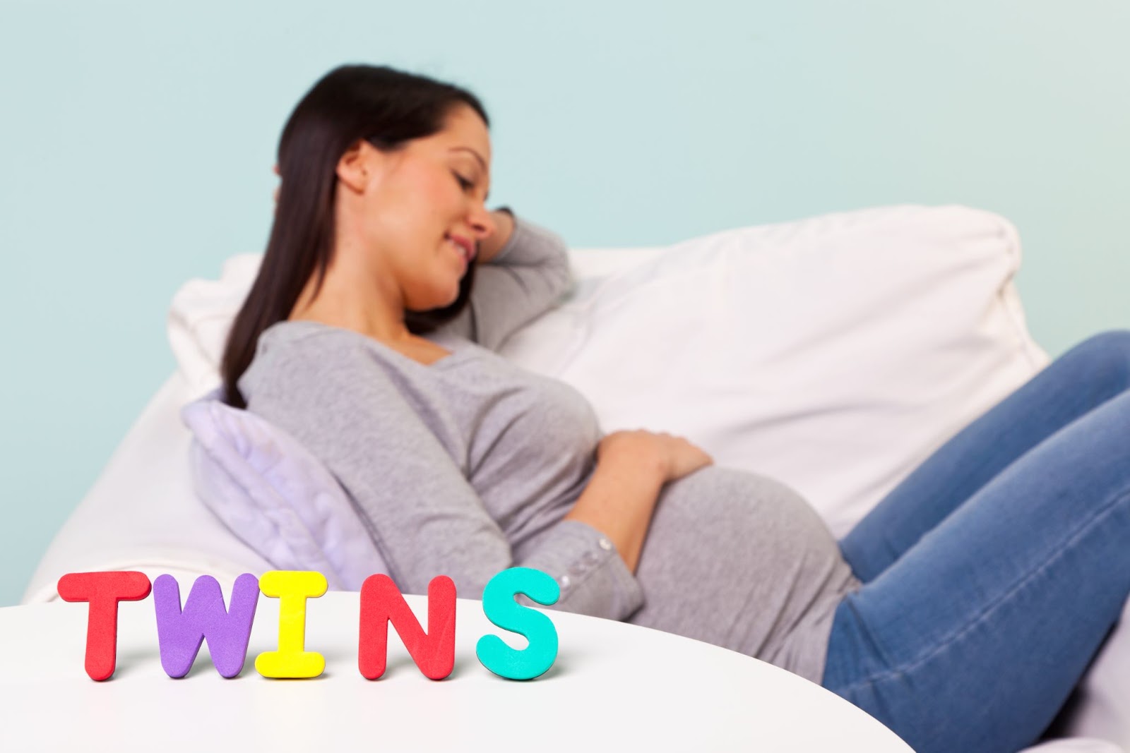Pregnant woman looks down at her belly and smiles behind letters spelling out “Twins”.